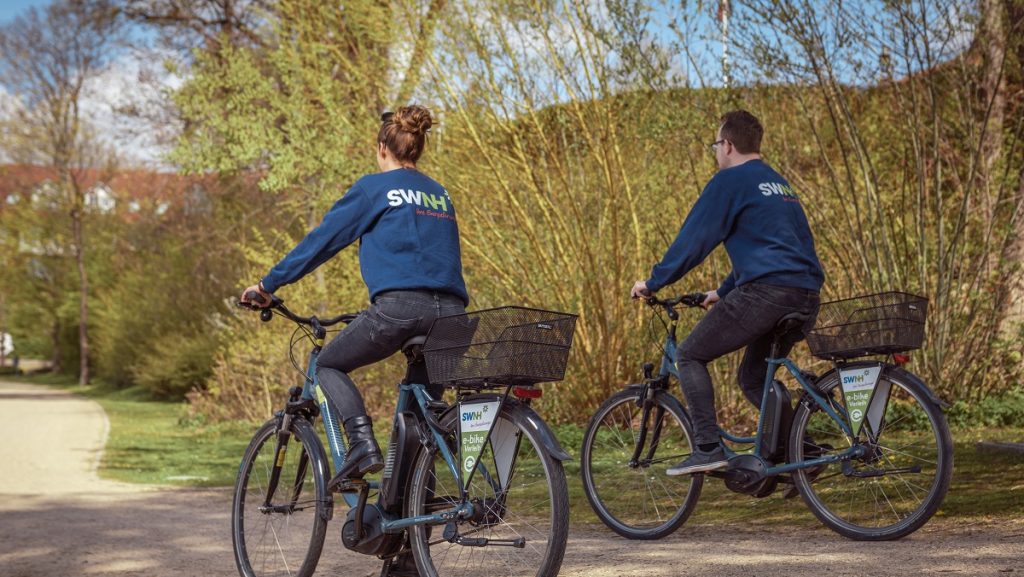 Zwei Personen fahren mit blauen E-Bikes auf einem Weg in einem Park mit Bäumen im Frühling