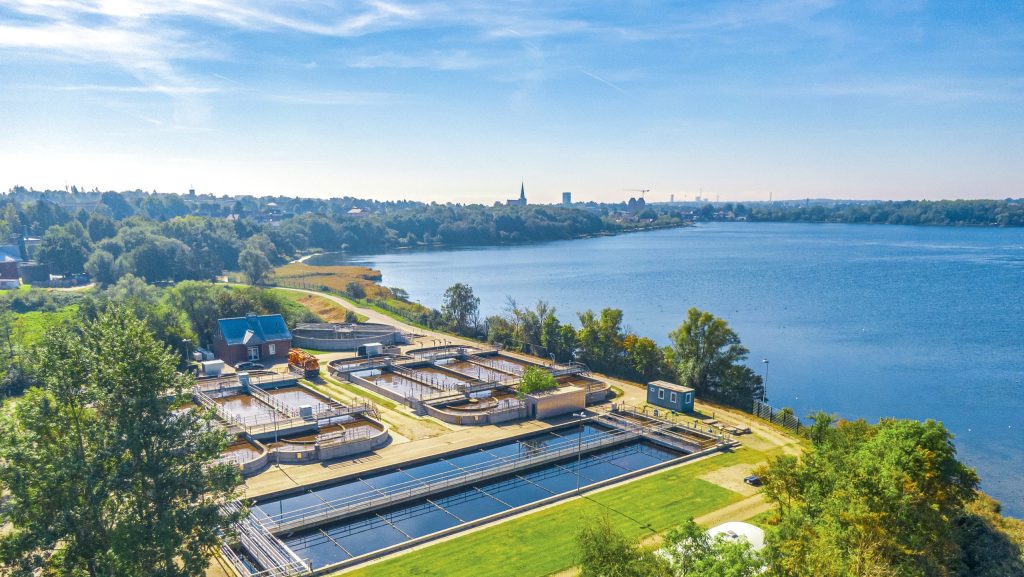 Kläranlage an einem Seeufer mit Bäumen und Stadt im Hintergrund unter blauem Himmel